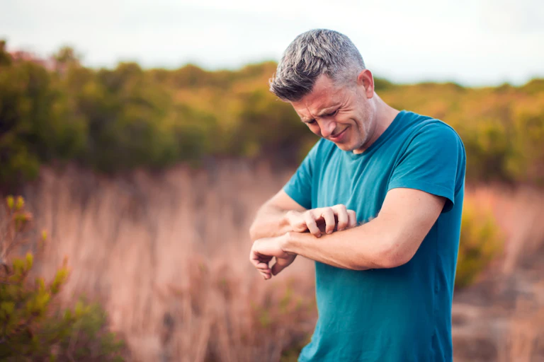 A man outdoors scratches his forearm.