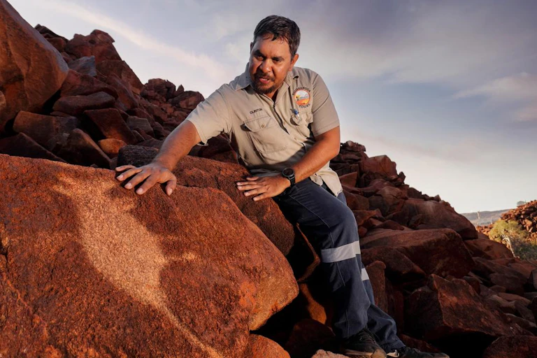 Man sitting on rocks and looking down and reaching towards at a rock art image.