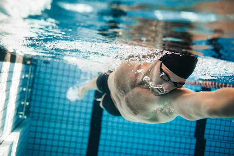 A male swimmer does the breast stoke in the pool. He's wearing goggles and a swim cap