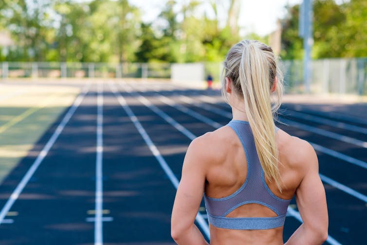 A woman stands on a track in a sports bra, you can see the muscles in her back.