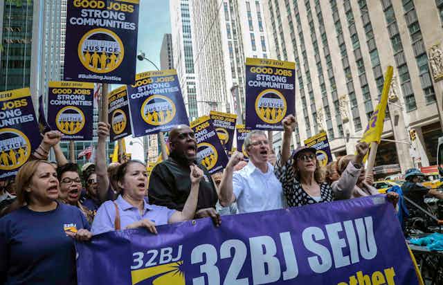 a diverse crowd carrying pro-union signs and a big banner hold up their fists while marching on a street surrounded by tall buildings