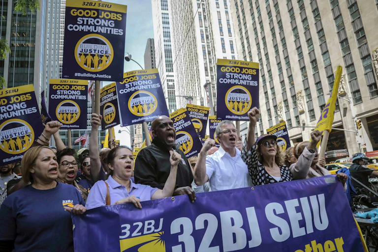 a diverse crowd carrying pro-union signs and a big banner hold up their fists while marching on a street surrounded by tall buildings