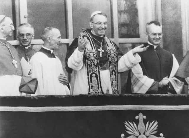 White male priests, including one with a large cross necklace, smile as they watch a crowd.
