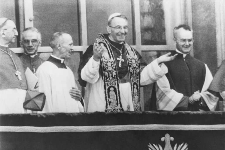 White male priests, including one with a large cross necklace, smile as they watch a crowd.