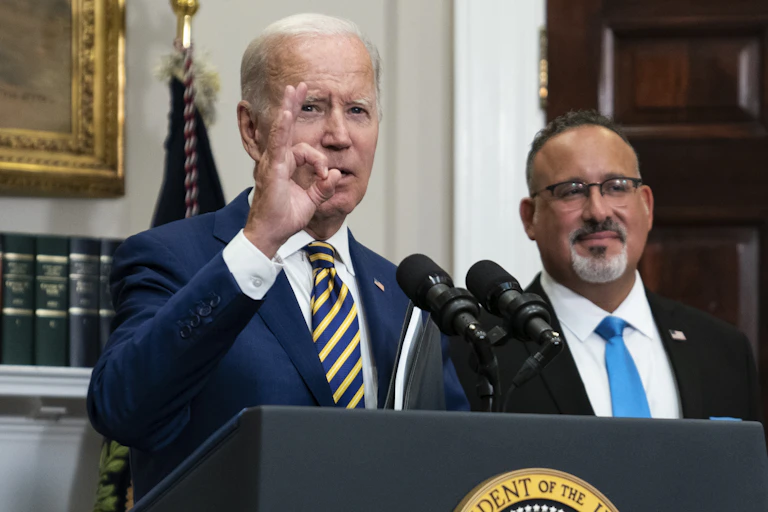 An elderly white man shapes his right hand into a 'zero' shape as he stands before a lectern