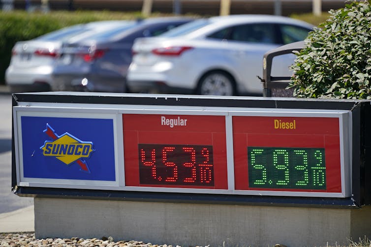 A gas station sign shows the price of gasoline with parked cars in background.