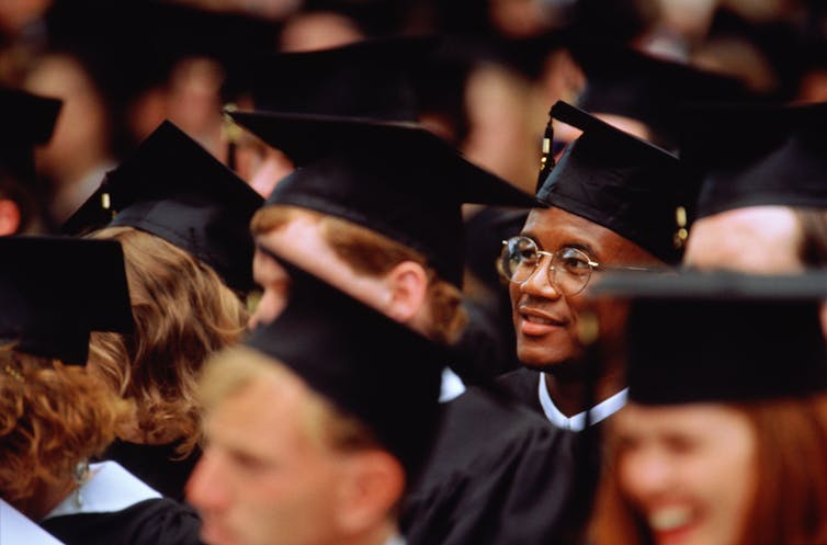 A black student with glasses wearing a cap in gown is the focus amid a sea of other faces.