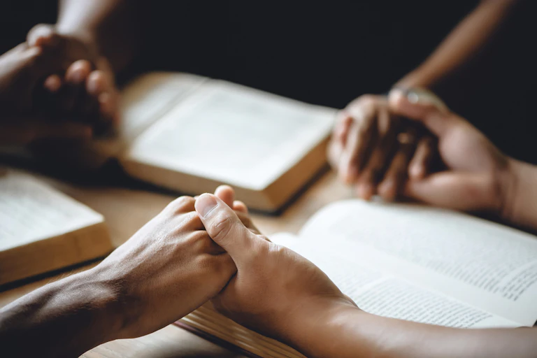 Three people hold hands as they sit around a table with open books.