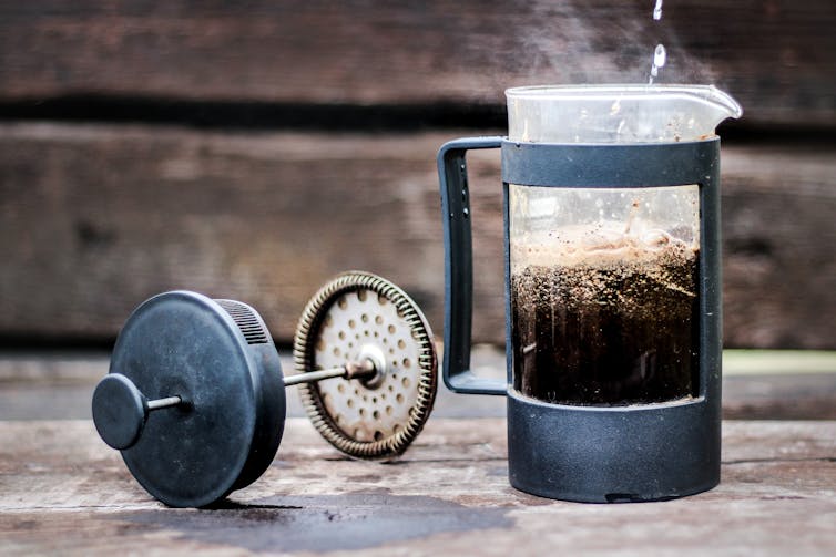 A glass beaker in a dark plastic frame with coffee steeping inside, the plunger laid next to it
