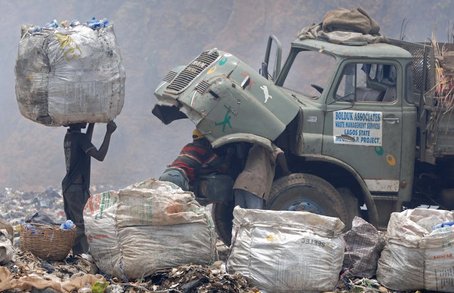 Man carries a bag of rubbish on his head watching men repairing a truck at a refuse dump site.