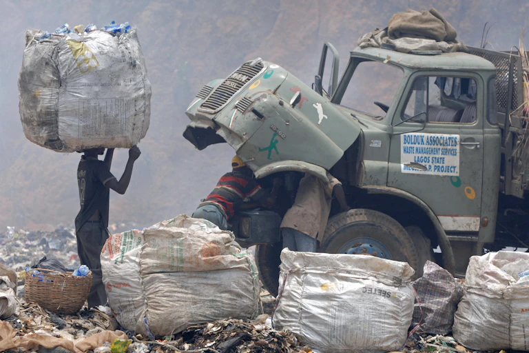 Man carries a bag of rubbish on his head watching men repairing a truck at a refuse dump site.