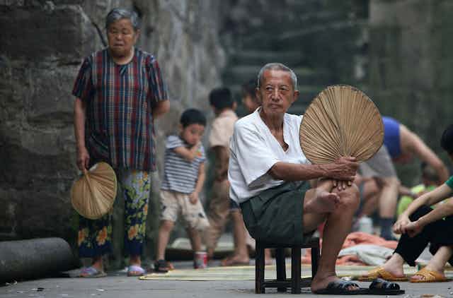 An older man sits with a woven fan.