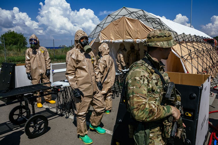 A soldier stands in the foreground as a half dozen people in hazmat suits and gas masks stand near stretchers outside a large tent