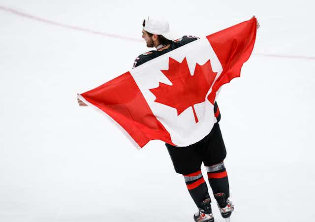 A man skates on an ice rink while carrying the Canadian flag behind his back.