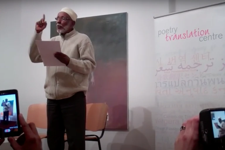 A man with a grey beard stands reading from a sheet of paper, a hand raised with a pointed finger. In the foreground people record him on their cellphones. fr