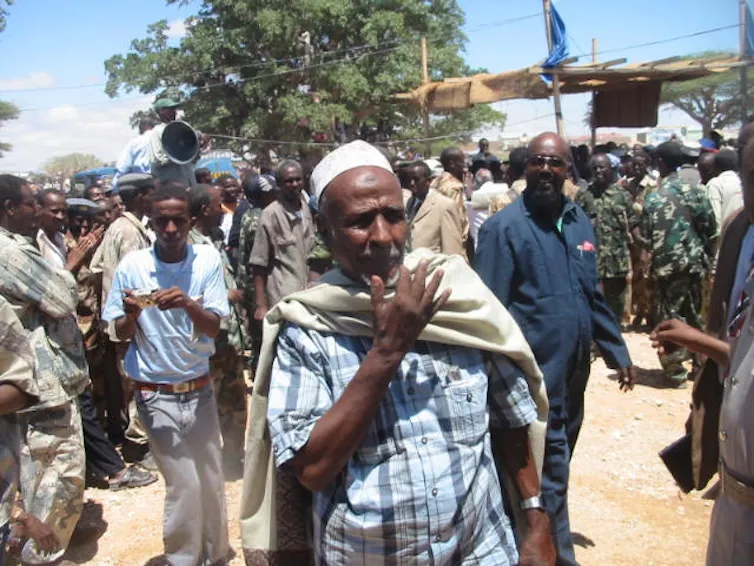 A man raises an arm as he speaks emphatically, standing outside with a small crowd of people in the background.