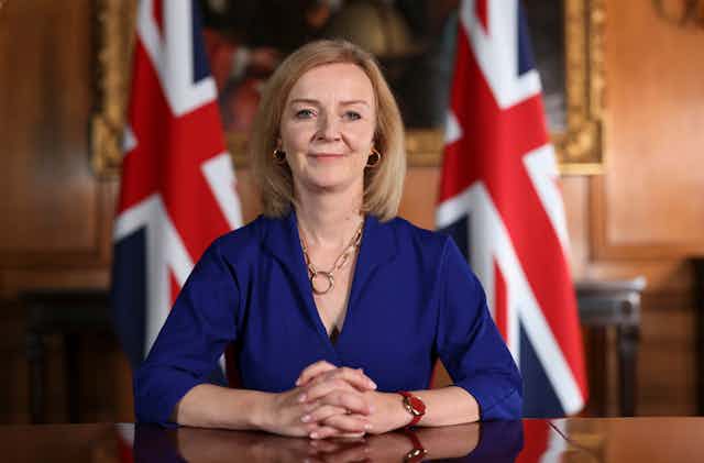 Ministerial portrait of Liz Truss sitting at a formal desk with her hands clasped on the desk, two UK flags behind her