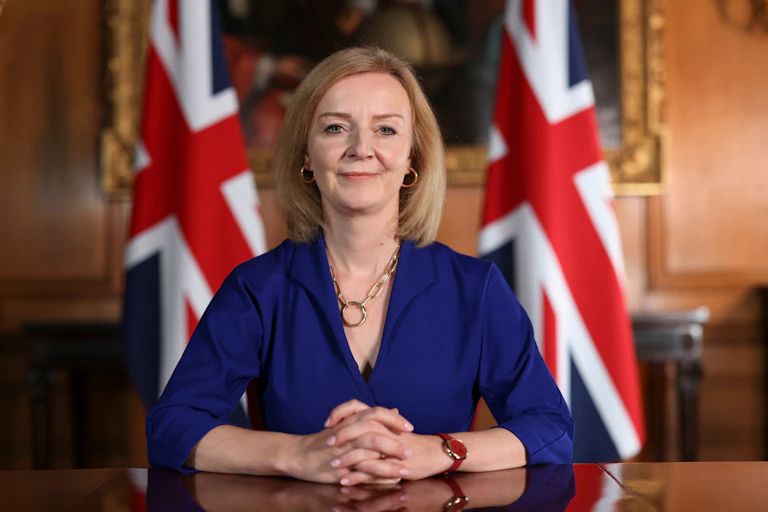 Ministerial portrait of Liz Truss sitting at a formal desk with her hands clasped on the desk, two UK flags behind her