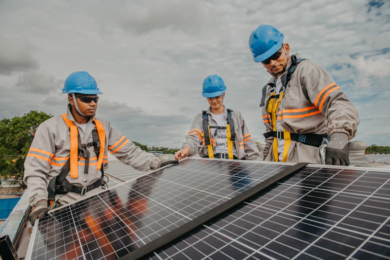Three people wearing aqua blue hardhats install a solar panel on a partly cloudy day.