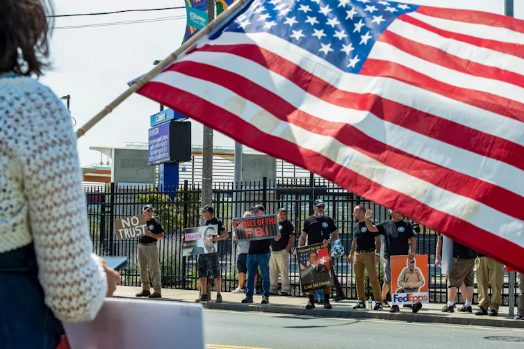 A person is seen waving a large American flag, across the street from people carrying signs that say 'treason,' and 'crimes of the FBI.'