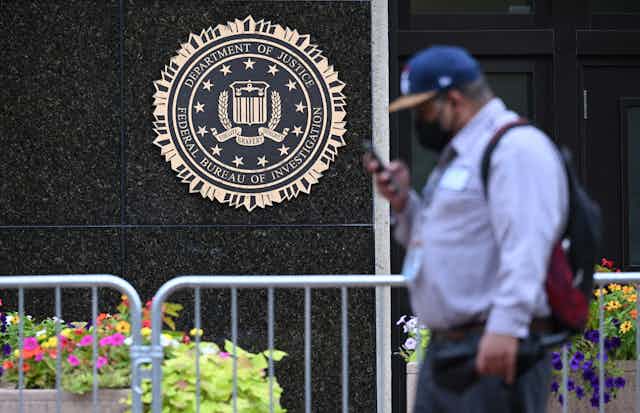A man in a blue shirt and blue cap reading his cellphone walks past a granite wall on which is affixed the seal of the Department of Justice and the FBI.