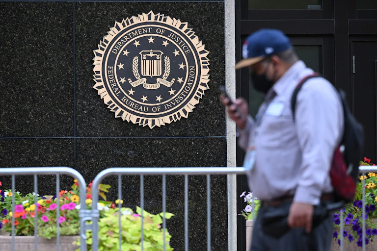 A man in a blue shirt and blue cap reading his cellphone walks past a granite wall on which is affixed the seal of the Department of Justice and the FBI.