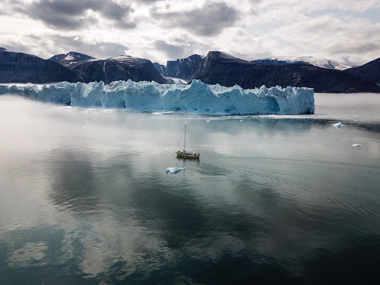 A tall ship with an even bigger iceberg behind it and a glacier in the distance.