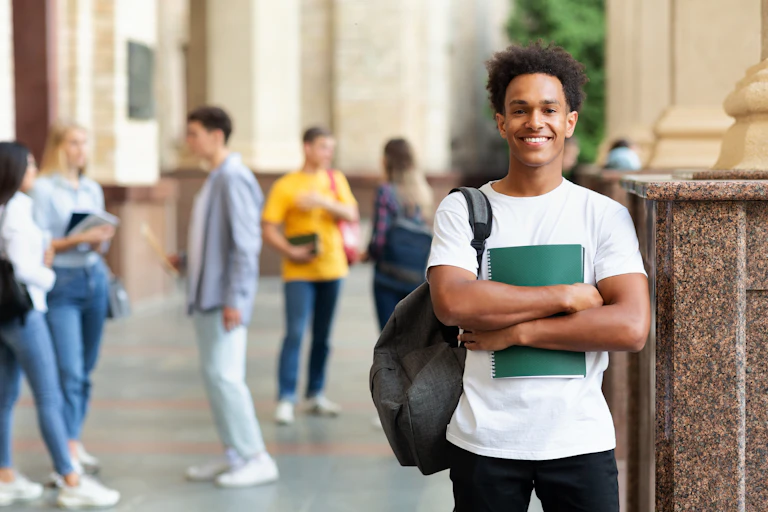 A young man with a backpack holding a notebook in the foreground smiling with other young people out of focus in the background