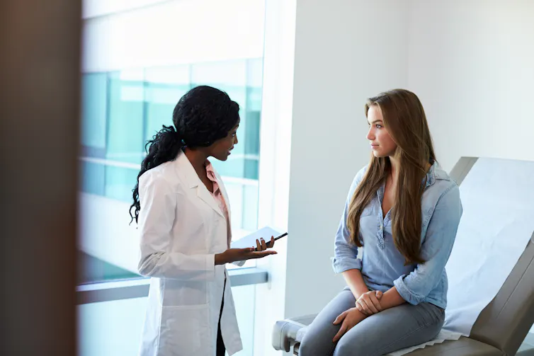 A young woman sitting on an exam table talking to a doctor in a white coat.