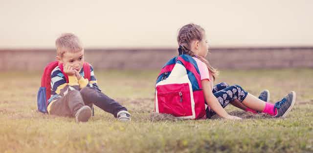 Dos escolares con mochila, niño y niña, sentados en la hierba con cara de hastío. 
