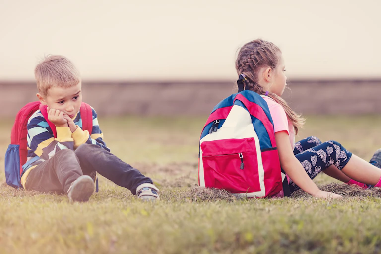 Dos escolares con mochila, niño y niña, sentados en la hierba con cara de hastío.