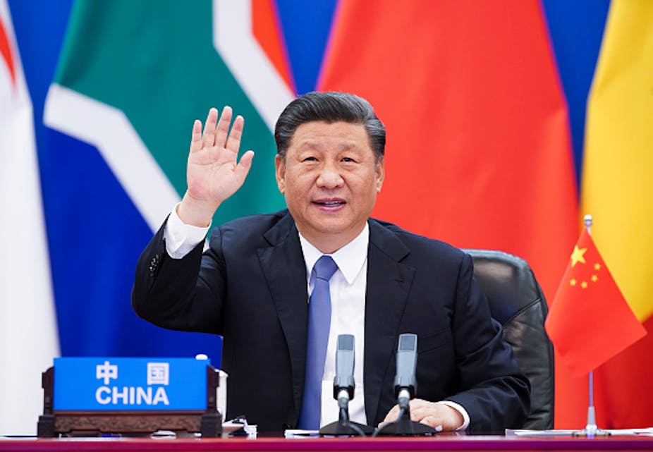A man in suit and tie sits in front of flags with his right hand raised and a sign reading "China" in front of him