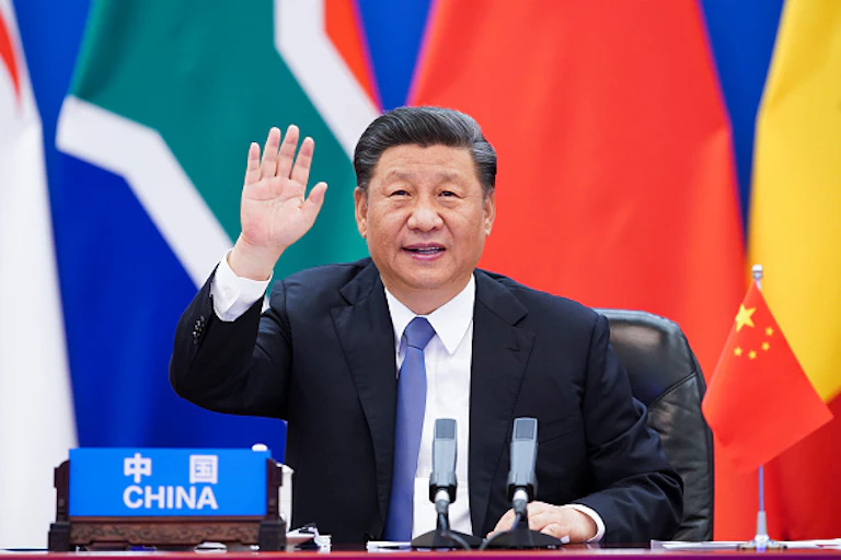 A man in suit and tie sits in front of flags with his right hand raised and a sign reading "China" in front of him