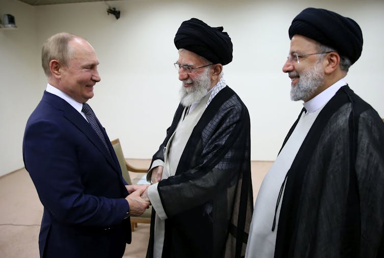 Three men greet one another -- one in a black suit, the other two in black clerical robes with black headwraps