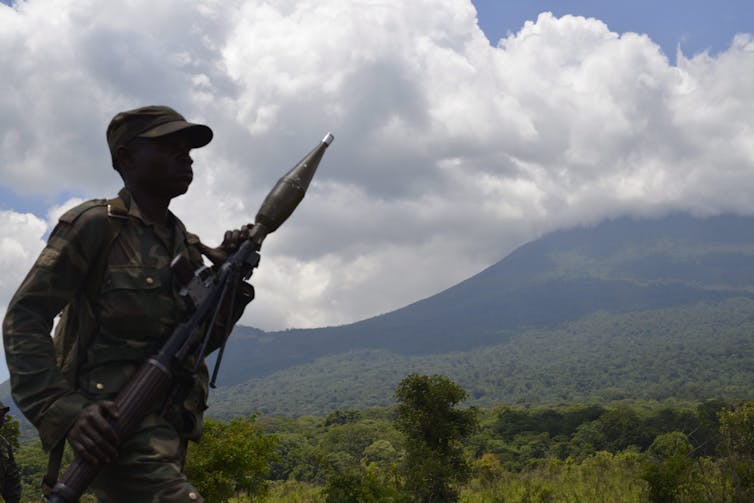 A soldier carrying a large weapon is seen against a backdrop of green mountains and clouds.
