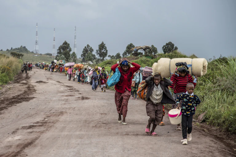 A line of people, a young boy in front, walk along a dusty road carrying their belongings.