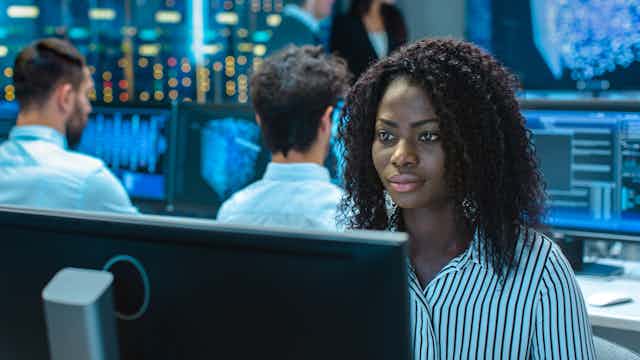 A dark-skinned woman in a striped blue shirt sits in front of a large computer monitor. There are two people behind her, backs to the camera, also looking at monitors whose contents are blurred.