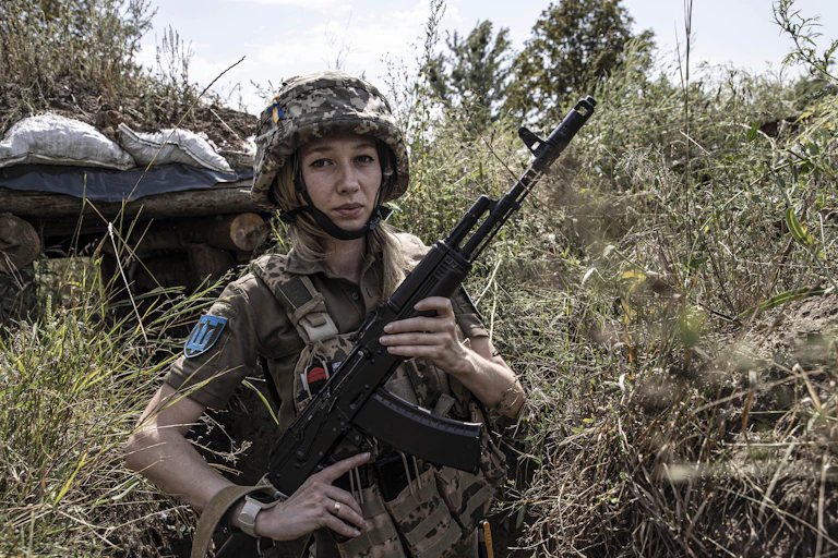 A young woman with light hair holds up a gun and wears camouflage military clothing, standing in a grassy area outside. She looks straight at the camera.