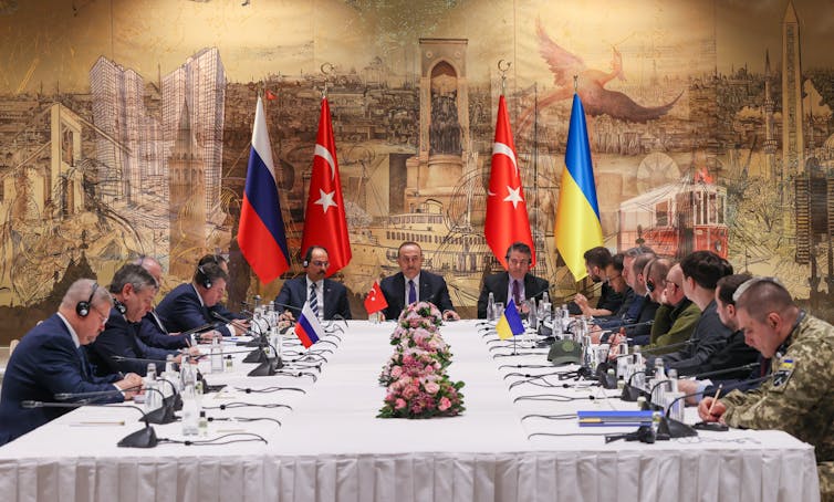 Two rows of men in dark suits sit around a formal white table, with three men sitting at the head of it. Behind them are Ukrainian, Turkish and Russian flags.
