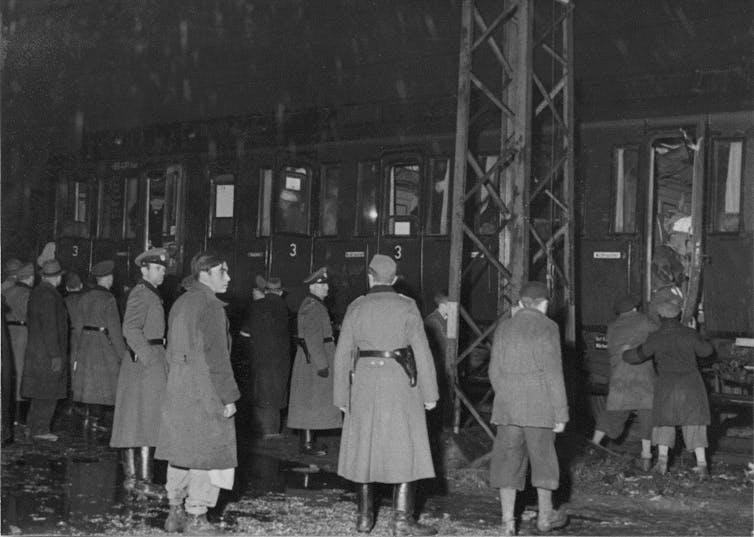 Soldiers watching a train filled with people as a person is pushed onto it.