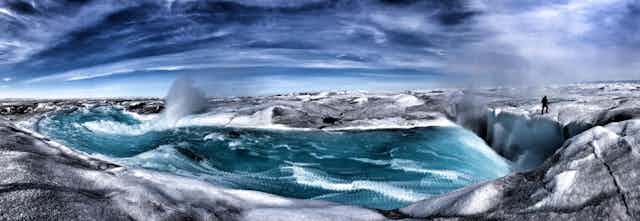 Alun Hubbard stands at the edge of a crevasse where meltwater is cascading in. The pano style photo creates an effect that makes it look like a painting.