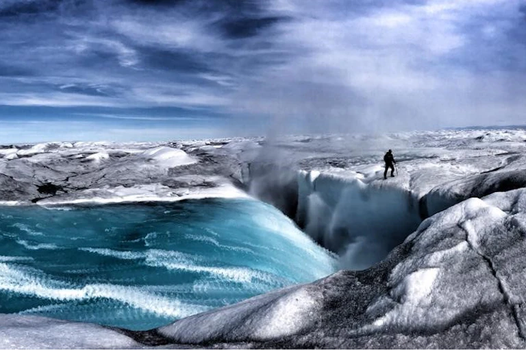 Alun Hubbard stands at the edge of a crevasse where meltwater is cascading in. The pano style photo creates an effect that makes it look like a painting.