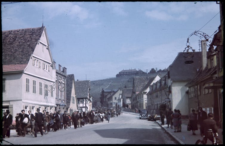 Hundreds of people being marched down a village street, while onlookers watch.