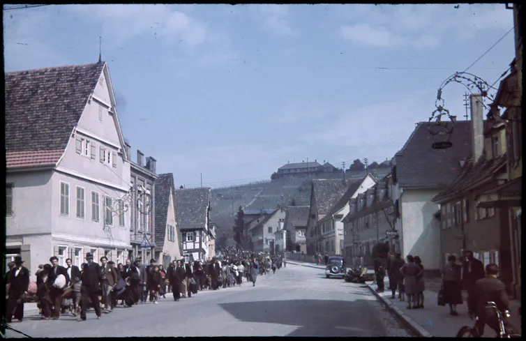 Hundreds of people being marched down a village street, while onlookers watch.