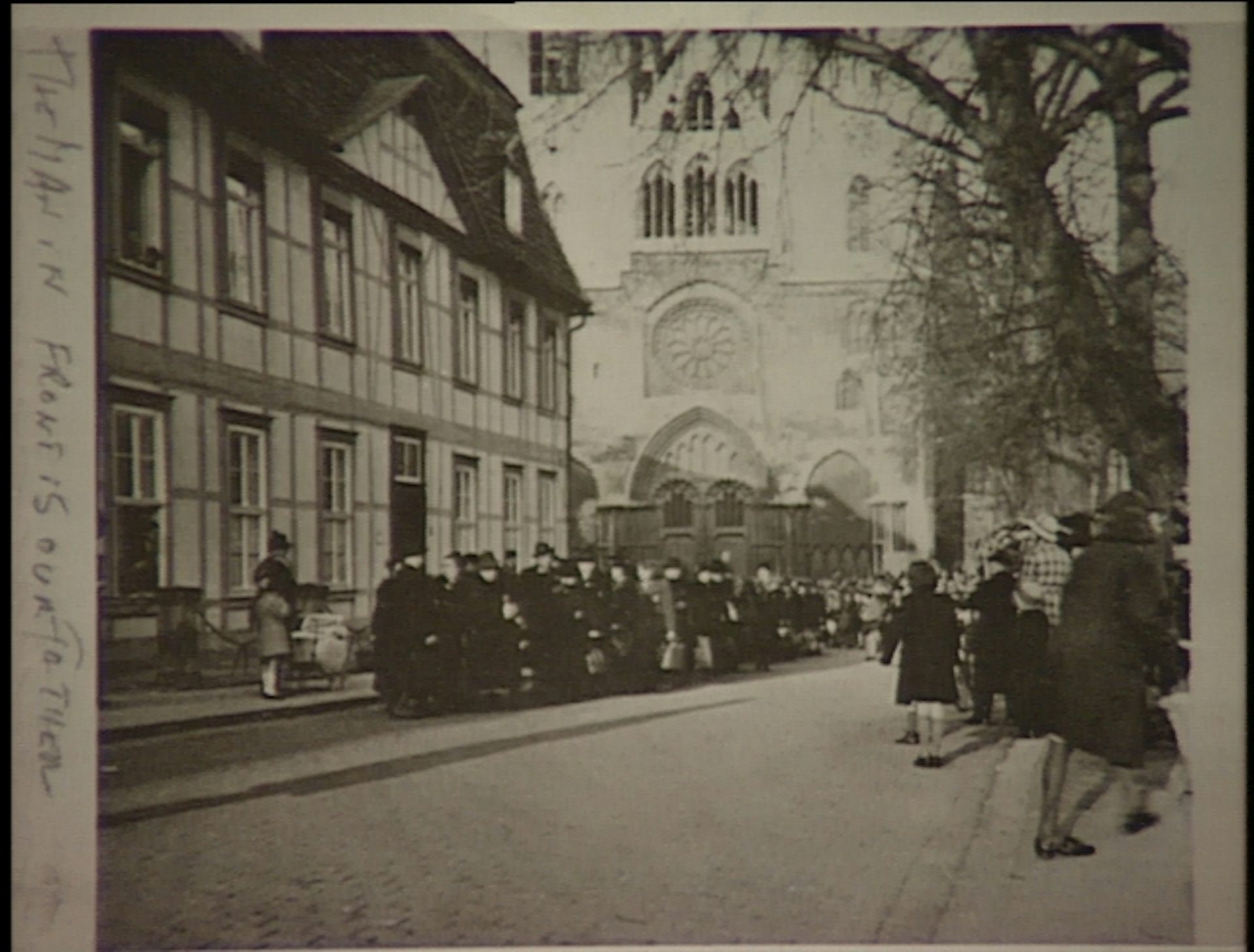 A large group of people assembled on the street in front of a timbered building and a large church, with people watching them on the other side of the street.