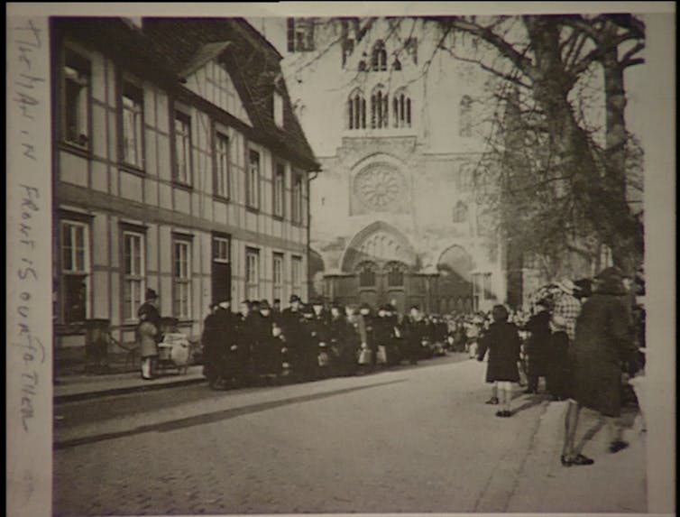 A large group of people assembled on the street in front of a timbered building and a large church, with people watching them on the other side of the street.