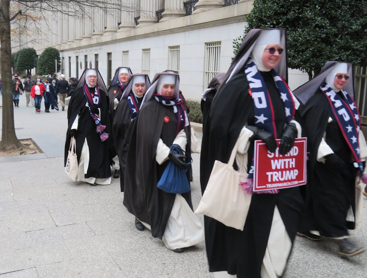 A group of nuns wearing full habits march, also wearing scarves that say 'Trump' and carrying a sign saying 'Stand with Trump.'