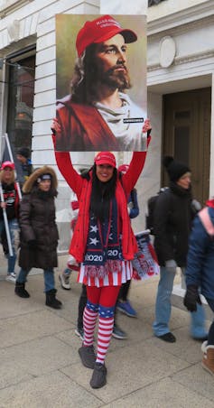 A woman holds a poster showing a portrait of Jesus wearing a red 'MAGA' ballcap.