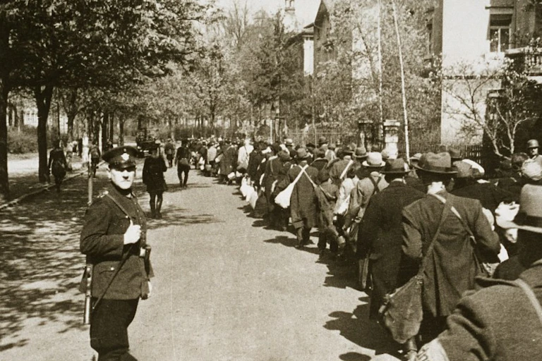 A large crowd of people carrying personal belongings in bundles and suitcases, watched over by a soldier.