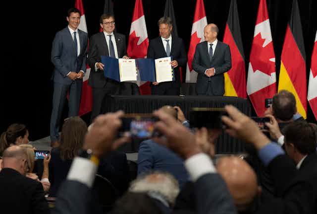 Four men in suits stand before a crown of photographers. Two of them are holding signed agreements with the Canadian and German flags visible on them.